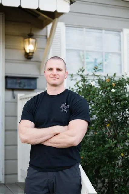 Man in black t-shirt with logo, smiles in front of a house, standing on porch with hand on railing.