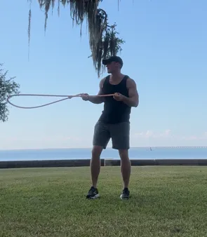 Man exercising with a resistance band outdoors. He is standing on grass, by the water, holding the band.